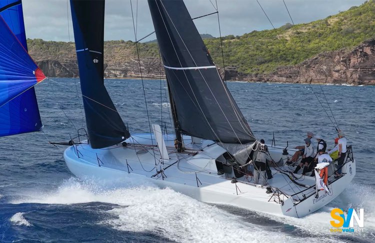 Vista di tre quarti: il piano velico e la coperta essenziale accompagnano una carena offshore dove la prua scow è il dettaglio più evidente