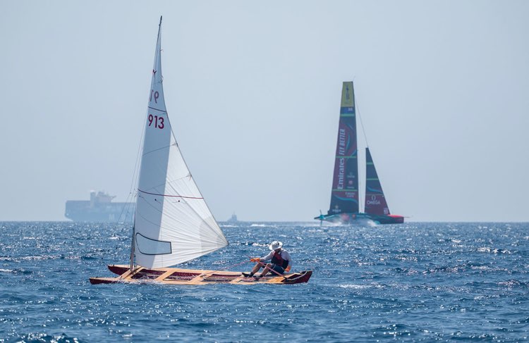 Un Patin a Vela naviga con una AC75 di Coppa America sulla sfondo. Il minimo della tecnologia e poco più in la il suo massimo